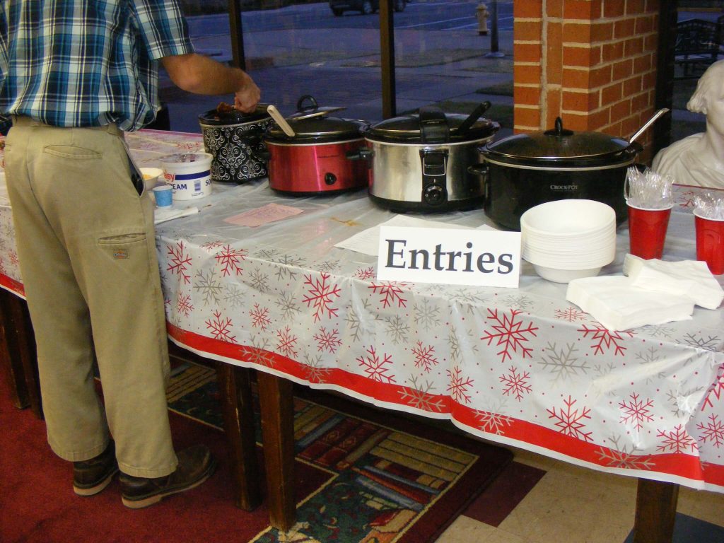 Four crock pots of Chili on the entries table with a man serving himself from one of the entries. 